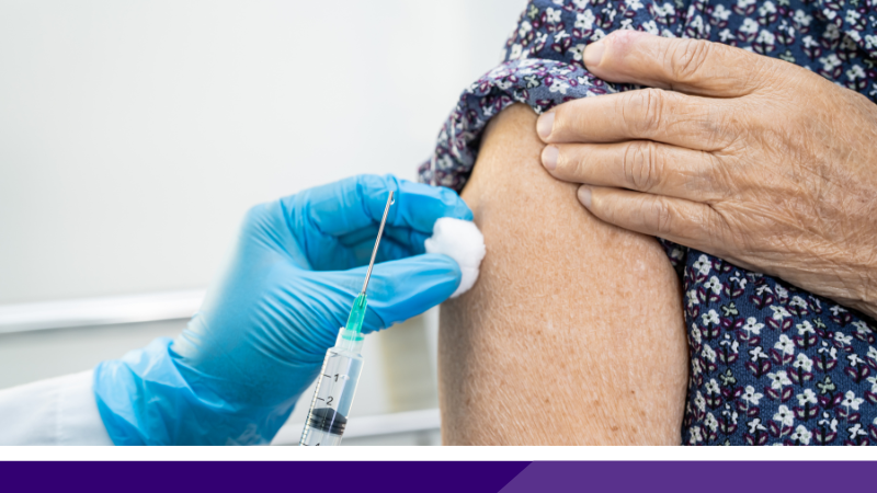 Close up of an older person's exposed shoulder, with a clinician's hand, wearing a blue rubber glove, wiping with a piece of cotton wool, while the other hand has a needile ready to deliver a vaccine.