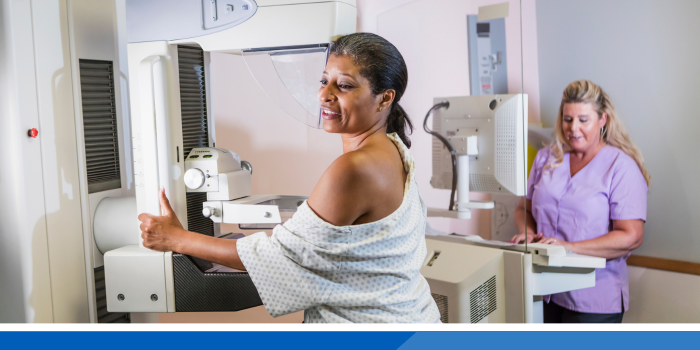 Lady in a hospital gown whilst having a mammogram. Nurse in the background stood behind a computer.