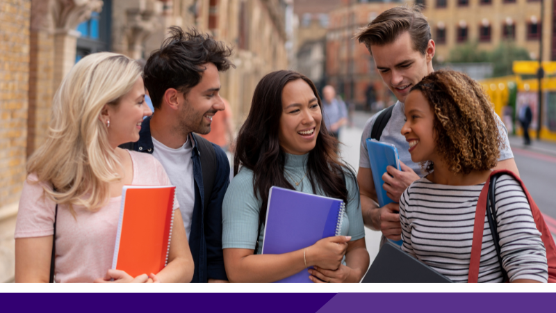 Five young people aged around 18 to 19 years old standing on the roadside in a town. A girl with long blonde hair holding a large orange notepad to her chest on the far left, next to a man with dark hair. To his left is a girl with long dark hair, holding a large purple notepad to her chest. A man with dark hair holding a light blue notepad is next to her while a black girl with shoulder length brown hair and a white stripey top and a red handbag over her shoulder is on the far right.