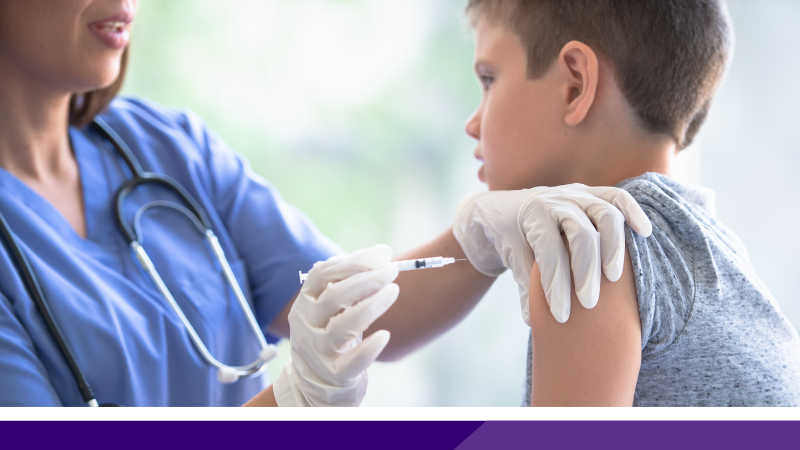 A young boy on the right is having an injection put into his left shoulder by a nurse wearing white latex gloves and a blue uniform.