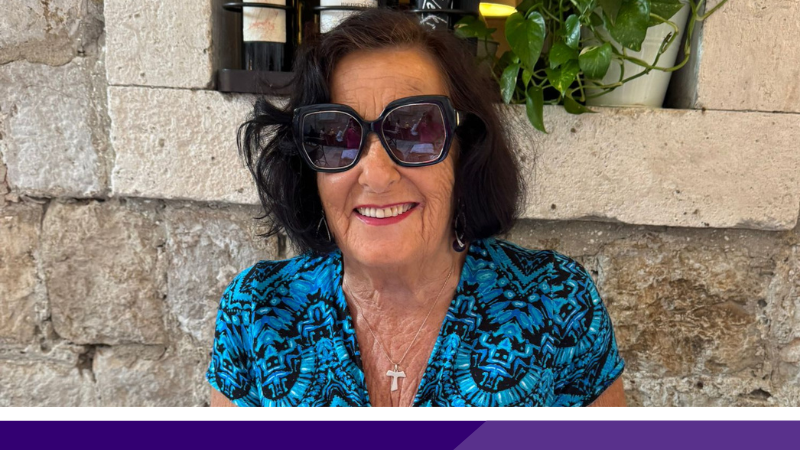 Head and shoulder of a woman in her 70s with shoulder-length black hair, wearing sunglasses and a turqoise and black dress, , sitting outside.