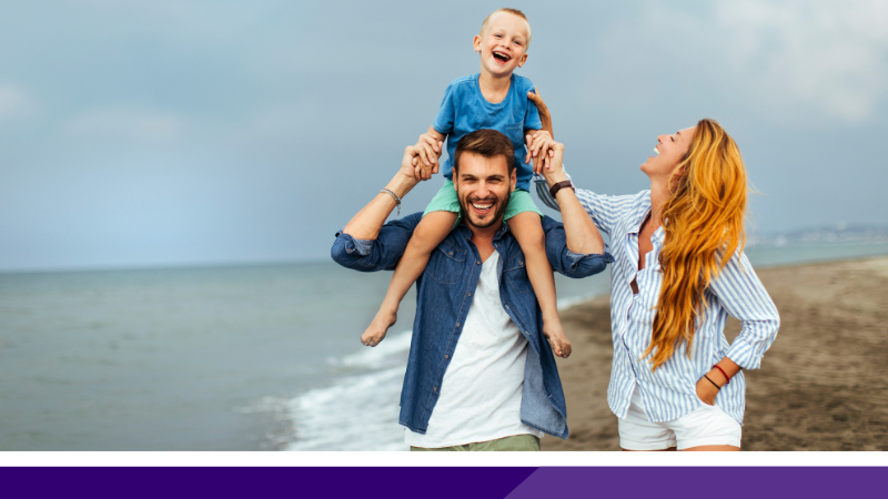 A father holds his son on his shoulders as they walk along the shore of a beach, with the wife walking alongside next to them.