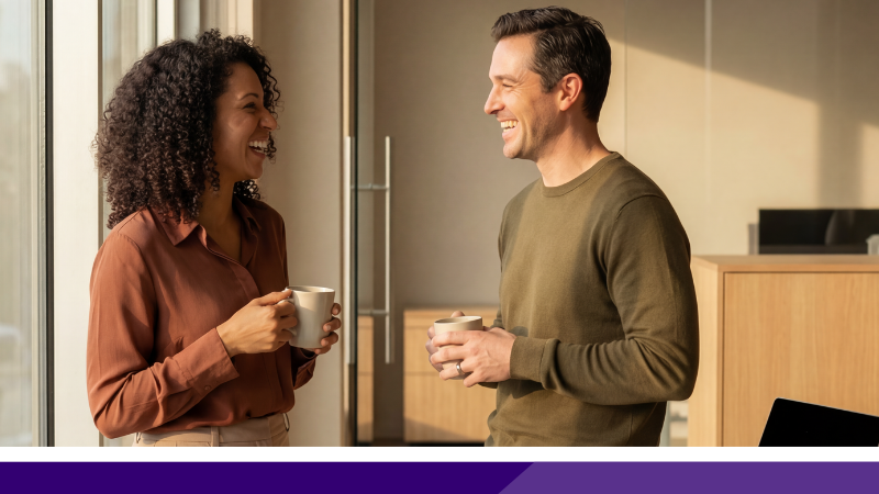A woman on the left with curly black hair holding a mug, smiling at a man with short brown hair, also holding a mug and smiling, in a light-coloured office space.