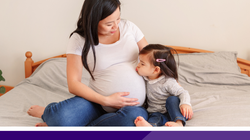 Pregnant woman sat on a bed next to a toddler who is kissing her belly
