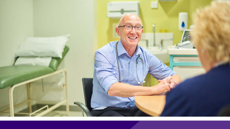 Doctor with a stethoscope round their neck talking to a patient. There is an examination couch in the background